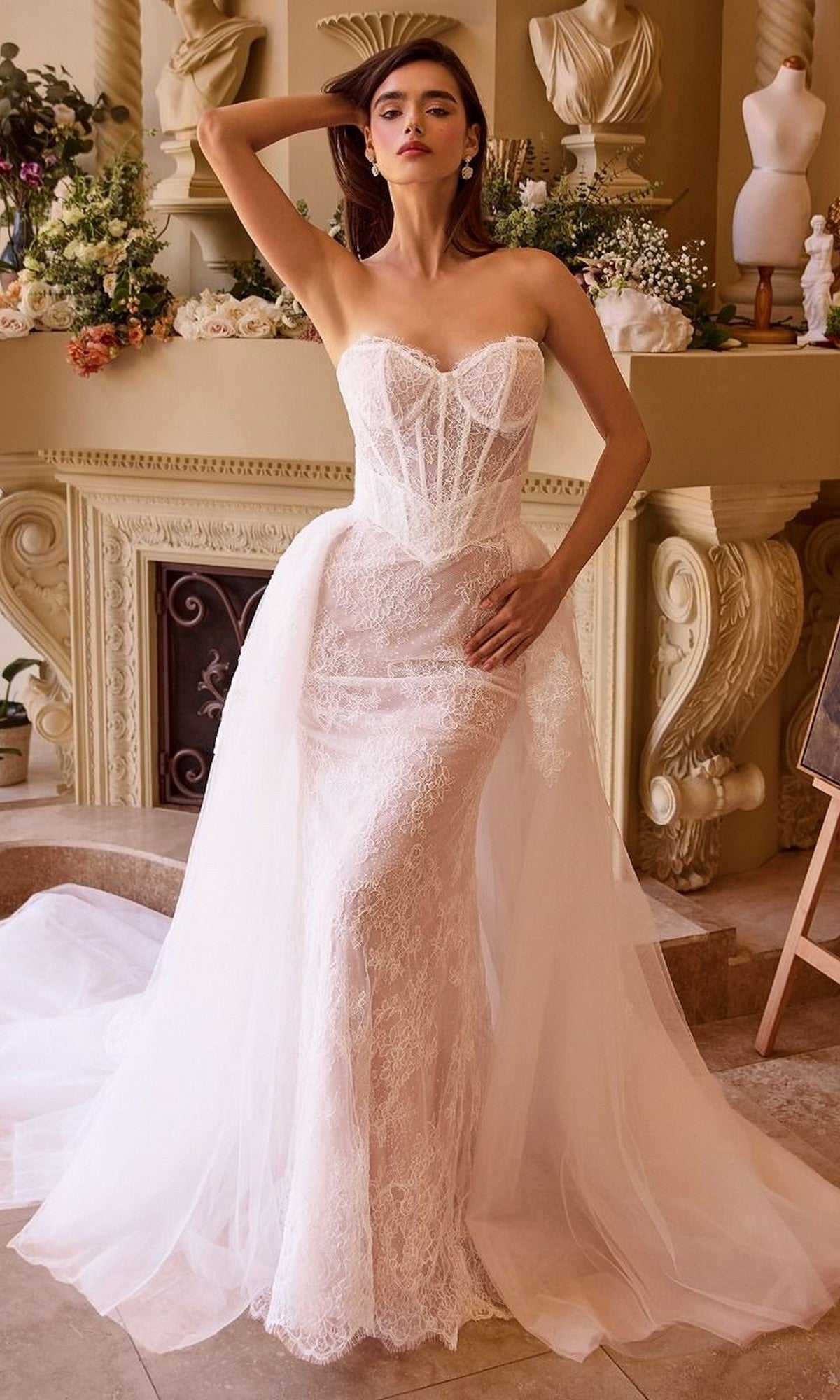 A woman wearing the Andrea and Leo WL044 Long Formal Dress in Off White/Nude, featuring a lace corset bodice with a sweetheart neckline, stands indoors before an ornate fireplace adorned with flowers and classical statues—an elegant choice for bridal celebrations.
